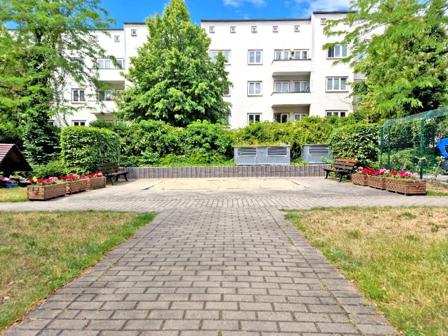 Innenhof Gepflasterter Weg zu einem Garten mit Blumenbeeten und einem Wohnhaus im Hintergrund.