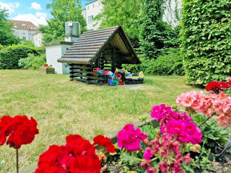 Innenhof Bunte Blumen im Vordergrund, im Hintergrund ein Spielhaus in einer grünen Wiese.