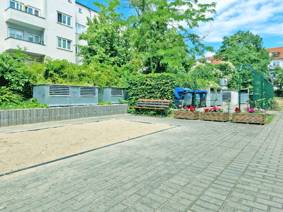 Innenhof Sandspielplatz mit Blumenbeeten und umgeben von Bäumen und Wohngebäuden.