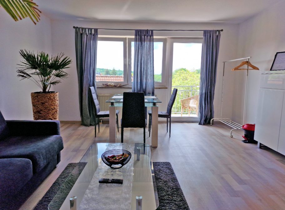 Wohnzimmer Heller Wohnraum mit einem Tisch, Stühlen, Sofa und großen Fenstern mit Blick ins Grüne.